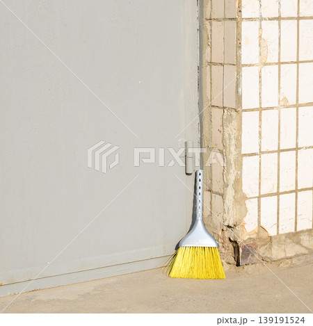 yellow broom with a gray handle stands on concrete at the base of a gray metal door in the sun yellow broom with a gray handle stands on concrete at the base of a gray metal door in the sun 139191524