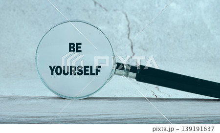 A magnifying glass reveals the important message Be Yourself clearly displayed on a light wooden table with a textured grey wall background 139191637