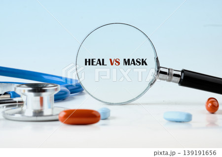 A magnifying glass reveals HEAL VS MASK written on a white surface. A blue stethoscope and several orange and blue pills surround the lens A magnifying glass reveals HEAL VS MASK written on a white surface. A blue stethoscope and several orange and blue pills surround the lens 139191656