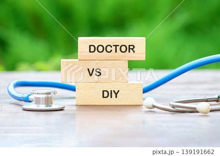 Stacked wooden blocks display Doctor VS DIY on a brown wooden surface. A blue stethoscope is nearby. Blurred green foliage forms the background Stacked wooden blocks display Doctor VS DIY on a brown wooden surface. A blue stethoscope is nearby. Blurred green foliage forms the background 139191662