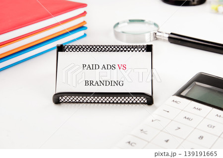 Paid Ads vs Branding on stand, white desk. Notebooks, calculator, magnifying glass for strategy Paid Ads vs Branding on stand, white desk. Notebooks, calculator, magnifying glass for strategy 139191665