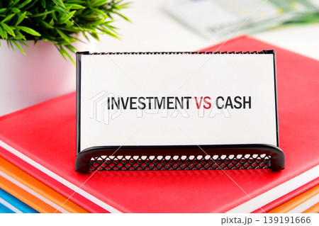 A white card displays Investment VS Cash in black and red text. It sits on colorful books in an office, symbolizing financial choices and planning A white card displays Investment VS Cash in black and red text. It sits on colorful books in an office, symbolizing financial choices and planning 139191666