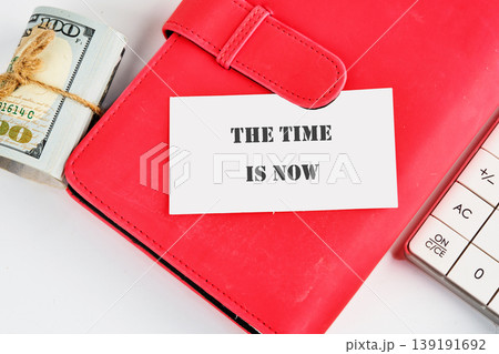 A red notebook, rolled US hundred-dollar bills tied with twine, and a calculator sit on a white surface, with a card reading The Time Is Now A red notebook, rolled US hundred-dollar bills tied with twine, and a calculator sit on a white surface, with a card reading The Time Is Now 139191692