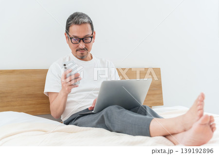 ベッドの上でスマートフォンとパソコンを使う中高年男性 139192896