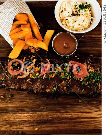 Grilled ribs garnished with diced green onions and red onion slices rest on a wooden platter. Potato fries, barbecue sauce, coleslaw 139193645