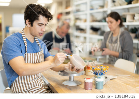 Young man applying coating with brush on ceramic piece 139194318
