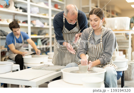 Eldery man helping young woman shape pottery on wheel Eldery man helping young woman shape pottery on wheel 139194320