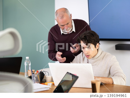 Mature male manager helping to guy during working at laptop in office Mature male manager helping to guy during working at laptop in office 139194961