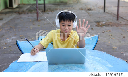 Cute Asian boy wearing headphones, waving hand to a laptop camera while studying online. Concept of virtual classroom, video conference, and digital communication for children's education. 139197174