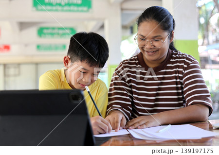 Happy Asian teenage girl or sister helping her younger brother with homework. They are using a digital tablet for e-learning at a wooden table. Concept of sibling support, tutoring, and education. Happy Asian teenage girl or sister helping her younger brother with homework. They are using a digital tablet for e-learning at a wooden table. Concept of sibling support, tutoring, and education. 139197175