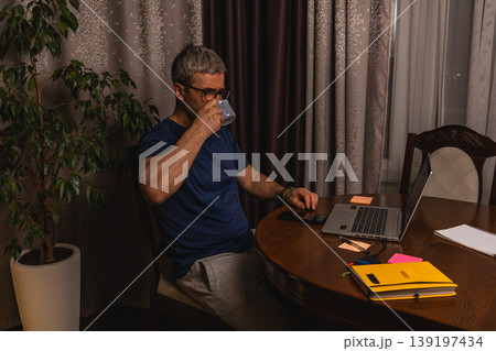 Tired freelancer drinking coffee while working late at night on laptop at home 139197434