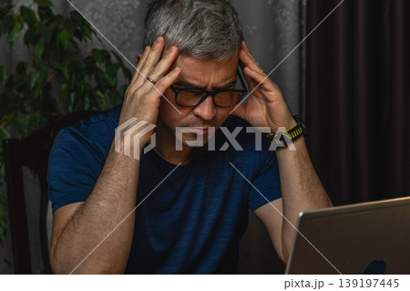 Overworked man holding head while working on laptop at night Overworked man holding head while working on laptop at night 139197445