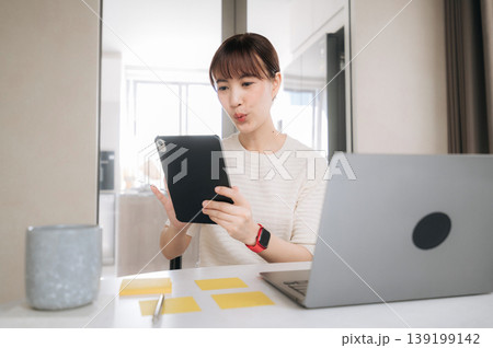 Asian woman doing remote business work from home focused on study with tablet and laptop on bright modern desk Asian woman doing remote business work from home focused on study with tablet and laptop on bright modern desk 139199142