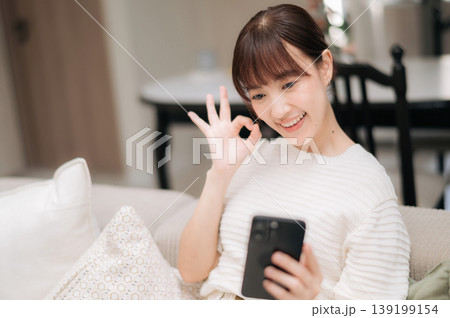 Happy asian woman doing remote work and business study at home showing ok hand sign using smartphone with joyful smile 139199154