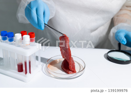 Scientist in protective gear holding lab-grown meat sample with tongs over petri dish in biotechnology lab. Cultured beef from in vitro cultivation process. Innovative sustainable protein production Scientist in protective gear holding lab-grown meat sample with tongs over petri dish in biotechnology lab. Cultured beef from in vitro cultivation process. Innovative sustainable protein production 139199976