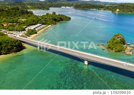 古宇利大橋とエメラルドグリーンの海に浮かぶ小島を真夏の空から捉えた写真 139200010