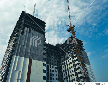 Low angle view of a high-rise building under construction with tower crane reaching into blue sky, modern concrete structure rising, urban development concept, architecture progress and industrial 139200373
