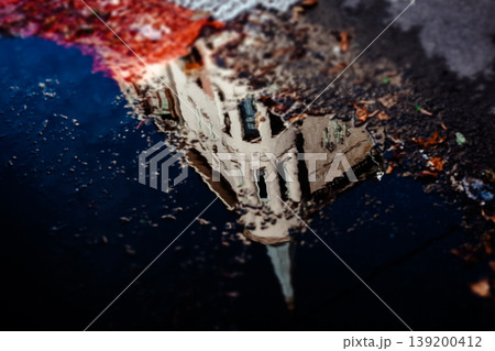 A striking reflection of a unique, old urban building in a puddle after rain Moody high-contrast image captures architectural details and the sky in the water's surface, highlighted by a splash of red A striking reflection of a unique, old urban building in a puddle after rain Moody high-contrast image captures architectural details and the sky in the water's surface, highlighted by a splash of red 139200412
