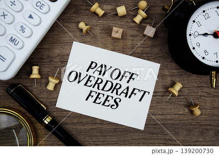 Pay Off Overdraft Fees text written on notepad with pen, alarm clock, magnifying glass and calculator on wooden background 139200730