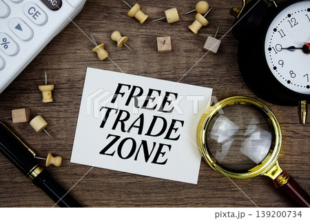 Free Trade Zone text written on notepad with pen, alarm clock, magnifying glass and calculator on wooden background 139200734