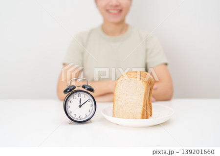 Happy smiling woman with whole wheat bread and alarm clock. concept of intermittent fasting, IF, meal timing, and choosing healthy carbohydrates for a balanced diet and weight loss management Happy smiling woman with whole wheat bread and alarm clock. concept of intermittent fasting, IF, meal timing, and choosing healthy carbohydrates for a balanced diet and weight loss management 139201660