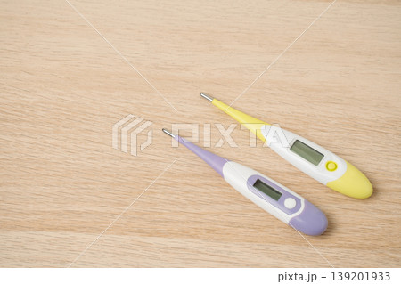 Two colorful digital thermometers rest on a light wooden surface, suggesting fever detection, home health care and illness monitoring. Clear LCD displays practical everyday temperature measurement. Two colorful digital thermometers rest on a light wooden surface, suggesting fever detection, home health care and illness monitoring. Clear LCD displays practical everyday temperature measurement. 139201933