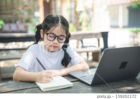 Young girl with glasses writes notes in a notebook while learning at an outdoor table near a laptop in a sunny garden setting 139202172