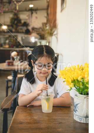 Young girl with glasses enjoying refreshing lemon drink in cozy cafe with beautiful yellow flowers on wooden table, perfect for lifestyle and family themes 139202449
