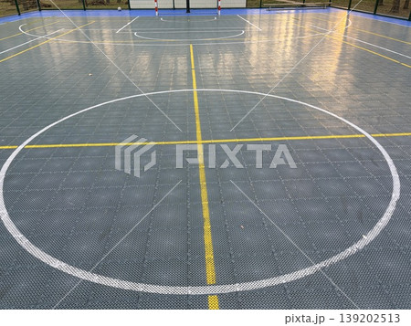 A modern small sports ground for basketball and football is surrounded by a high fence and is located in an open area 139202513
