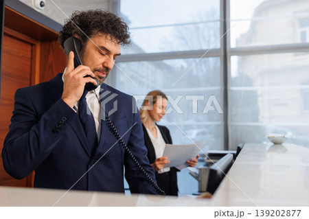 Receptionist talking on office phone with female employee in background Receptionist talking on office phone with female employee in background 139202877
