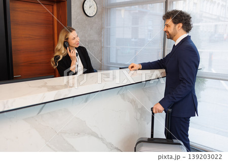 Professional Receptionist Speaking on Phone While Welcoming Business Guest at Hotel Front Desk Professional Receptionist Speaking on Phone While Welcoming Business Guest at Hotel Front Desk 139203022