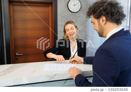 Receptionist Pointing at Contract While Guest Signs Paperwork at Modern Hotel Lobby Desk 139203030