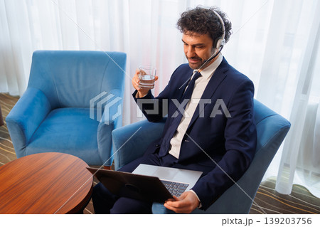 Smiling Businessman in Suit with Headset Using Laptop and Holding Glass of Water During Video Call Smiling Businessman in Suit with Headset Using Laptop and Holding Glass of Water During Video Call 139203756