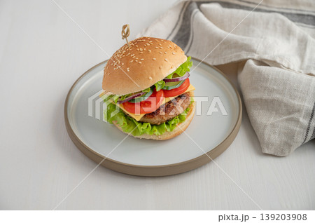 Beef burger with tomato, cheese, and lettuce on a plate on a light wooden background with napkin. Beef burger with tomato, cheese, and lettuce on a plate on a light wooden background with napkin. 139203908