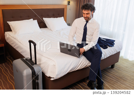 Business traveler working on laptop while sitting on hotel bed with suitcase Business traveler working on laptop while sitting on hotel bed with suitcase 139204582