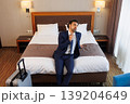 Tired Businessman Sitting on Hotel Bed After Business Trip with Suitcase in Modern Room 139204649