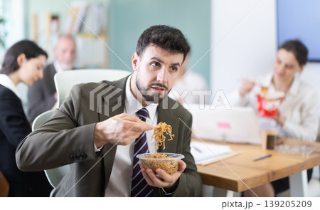 Young businessman having takeout meal during lunch break at work sitting at workplace at laptop with other colleagues in office 139205209