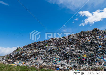 Large mound of waste piled in a garbage dump with multiple colors visible and birds flying overhead on a sunny day 139205792
