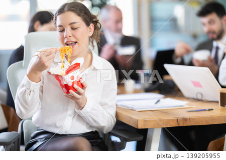 Young beautiful businesswoman having takeout meal during lunch break at work sitting at workplace at laptop with other colleagues in office Young beautiful businesswoman having takeout meal during lunch break at work sitting at workplace at laptop with other colleagues in office 139205955
