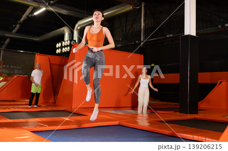 Enthusiastic woman leaping at vibrant trampoline gym with friends Enthusiastic woman leaping at vibrant trampoline gym with friends 139206215