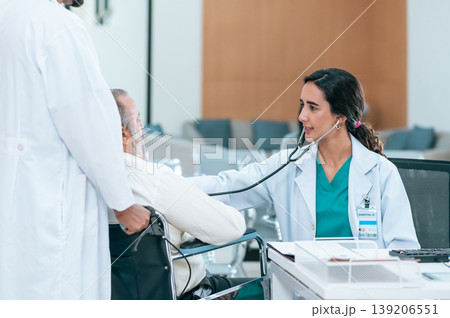 Old patient man sitting in wheelchair while young female doctor using stethoscope to check his heartbeat, elderly health checkup, happiness hospital. Old patient man sitting in wheelchair while young female doctor using stethoscope to check his heartbeat, elderly health checkup, happiness hospital. 139206551