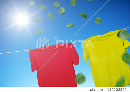 Vibrant red and yellow t-shirts hanging on a clothesline against a clear blue sky. Bright sun shines as fresh green leaves blow in the wind, representing freshness and clean laundry 139208262