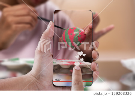 A close-up of hands holding a smartphone, viewing a live social media stream of an artist painting a decorative Easter egg. Features like heart reactions are visible on screen A close-up of hands holding a smartphone, viewing a live social media stream of an artist painting a decorative Easter egg. Features like heart reactions are visible on screen 139208326