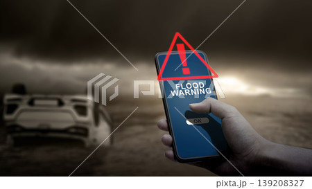 A hand holds a smartphone showing a flood warning alert, with an overturned car partially submerged in water under a dark, stormy sky in the background A hand holds a smartphone showing a flood warning alert, with an overturned car partially submerged in water under a dark, stormy sky in the background 139208327
