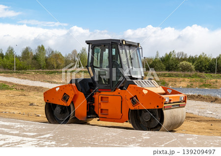 A roller is resting on a dirt road at a construction site near green trees on a sunny day with some clouds in the sky A roller is resting on a dirt road at a construction site near green trees on a sunny day with some clouds in the sky 139209497