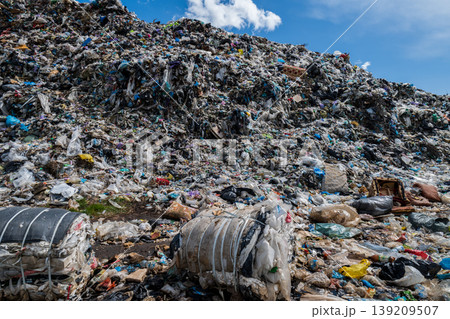 Large piles of trash cover a landfill site with visible plastic bags and debris scattered across the landscape in bright sunlight Large piles of trash cover a landfill site with visible plastic bags and debris scattered across the landscape in bright sunlight 139209507