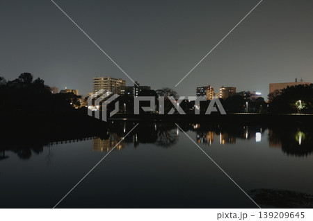暗く情緒的な空の下、低層ビルの微かな光を映し出す静かな湖の夜景 暗く情緒的な空の下、低層ビルの微かな光を映し出す静かな湖の夜景 139209645