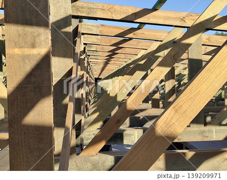 Wooden roof frame structure under construction with beams rafters and supports forming geometric pattern in sunlight at building site. Construction, carpentry, housing development Wooden roof frame structure under construction with beams rafters and supports forming geometric pattern in sunlight at building site. Construction, carpentry, housing development 139209971
