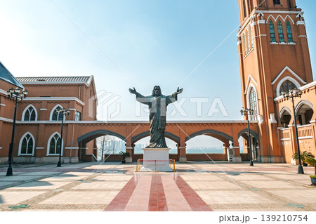 Jesus Christ statue with outstretched arms stands in a sunny plaza before a brick church, overlooking water and a distant city Jesus Christ statue with outstretched arms stands in a sunny plaza before a brick church, overlooking water and a distant city 139210754