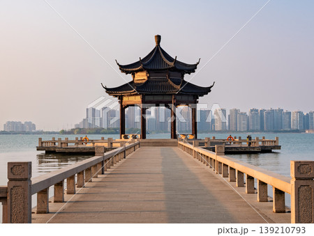 Ornate pavilion on serene lake connecting to modern cityscape in eastern China during golden hour Ornate pavilion on serene lake connecting to modern cityscape in eastern China during golden hour 139210793